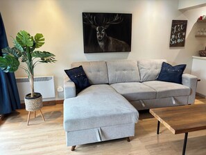 A living room with a grey sectional sofa, a wooden coffee table, a potted plant, and a deer painting on the wall.
