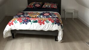 Une chambre avec un plafond en pente, un lit recouvert d’une courtepointe aux motifs floraux colorés, un luminaire suspendu blanc et une petite table de chevet.