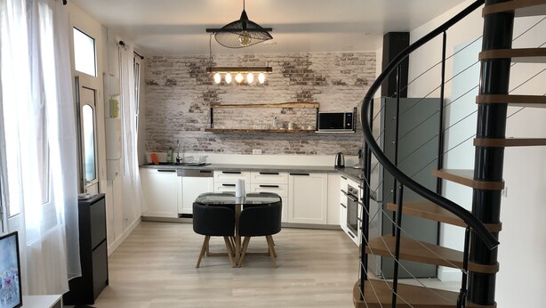 Une cuisine moderne avec des armoires blanches, un comptoir noir et un mur en briques. On y trouve un coin repas avec une table en verre et des chaises noires. Un escalier en colimaçon avec des marches en bois mène à un niveau supérieur.