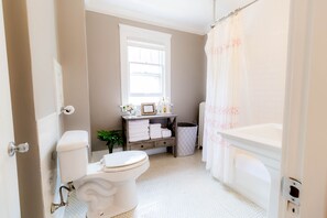 Et badeværelse med toilet, vask og badekar. Et vindue er placeret over vasken, og bruseforhænget har blomstermønster.