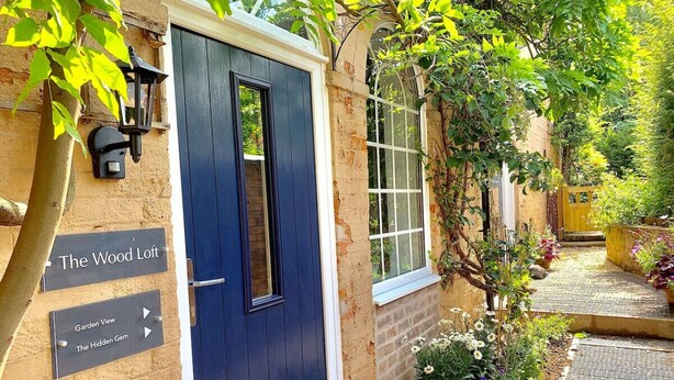 Uma porta azul escura com uma janela, rodeada por um jardim com diversas plantas e flores. Placas indicam "The Wood Loft" e "Garden View The Hidden Gem".