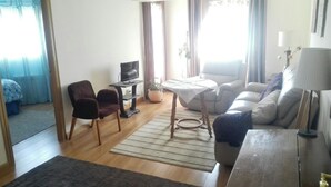 Uma sala de estar com sofá, cadeiras, televisão, uma mesinha e abajur.