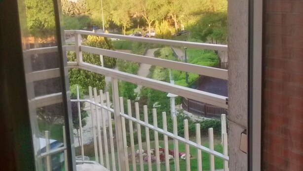 Uma varanda com guarda-corrimão branco, piso de azulejos e vista para uma paisagem verde com árvores e uma colina ao fundo.