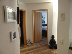 Un couloir doté d’un plancher en bois, une porte donnant sur une chambre à coucher et une statue de Bouddha posée sur le sol.