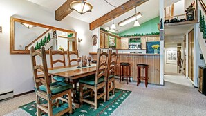 A dining area with a wooden table and chairs, a green rug, and a kitchen in the background. - Beaver Creek West 4bd/4ba free Ski shuttle The Perfect Mountain Nest (Avon)