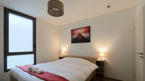 Un lit bien fait, avec une couverture rouge et des serviettes pliées, une table de chevet en bois avec une lampe, un tableau représentant un coucher de soleil accroché au mur et une grande fenêtre laissant entrer la lumière naturelle.