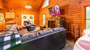 Ein Blockhaushaus-Interieur mit Kamin aus Stein, einem Ledersofa, einem an der Wand montierten Fernseher und einer Holzbalkendecke.