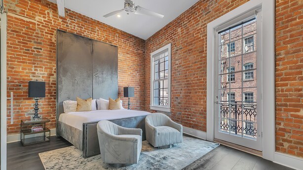 A bedroom with a brick wall, a large bed, two armchairs, a nightstand, and a window with a view of buildings.