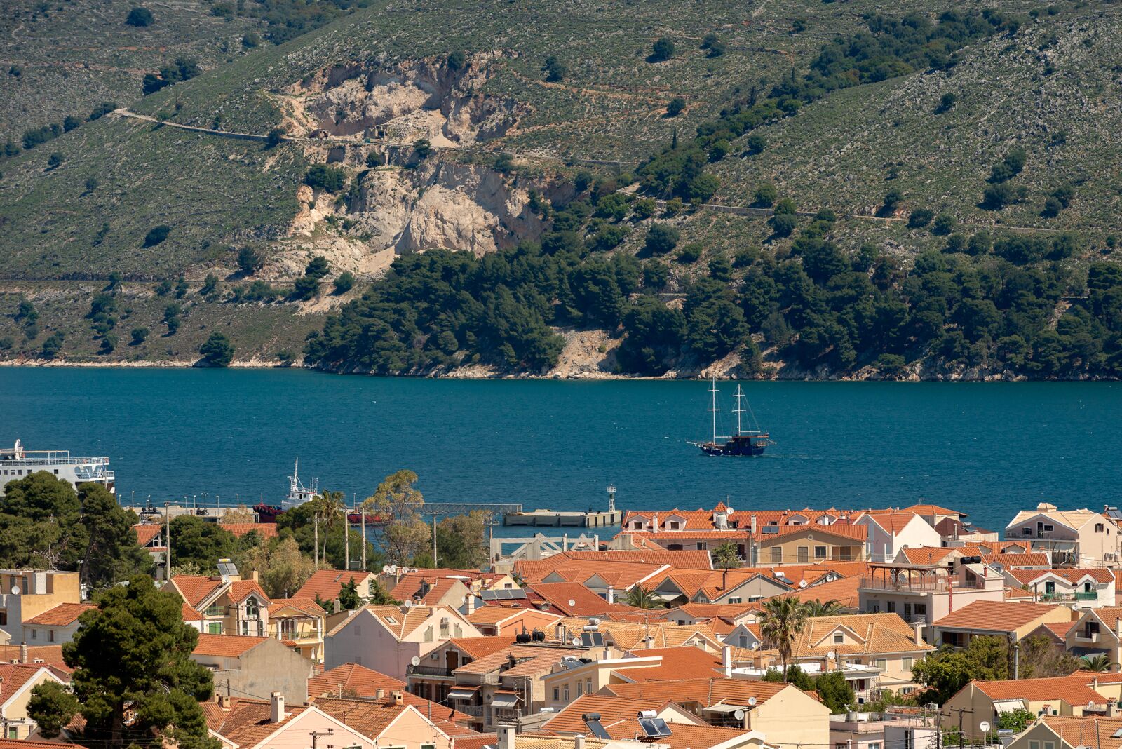 Une ville côtière aux maisons aux toits de tuiles rouges, un port de plaisance avec un voilier, et une colline rocheuse en arrière-plan.