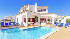 A poolside area with a clear blue pool, white buildings with red shutters, and blue lounge chairs.