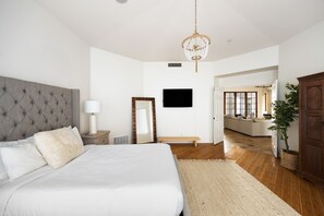 Une chambre avec un grand lit, une tête de lit en bois, une table de chevet avec une lampe, un téléviseur fixé au mur et une vue sur une autre pièce où se trouvent un banc et une plante.