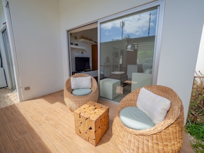 A modern outdoor seating area with wicker chairs and a wooden cube table, a glass door leading to an interior space with a TV, and a clear view of the sky. - Jaco Beach Condo Vereda B2 (Jacó)