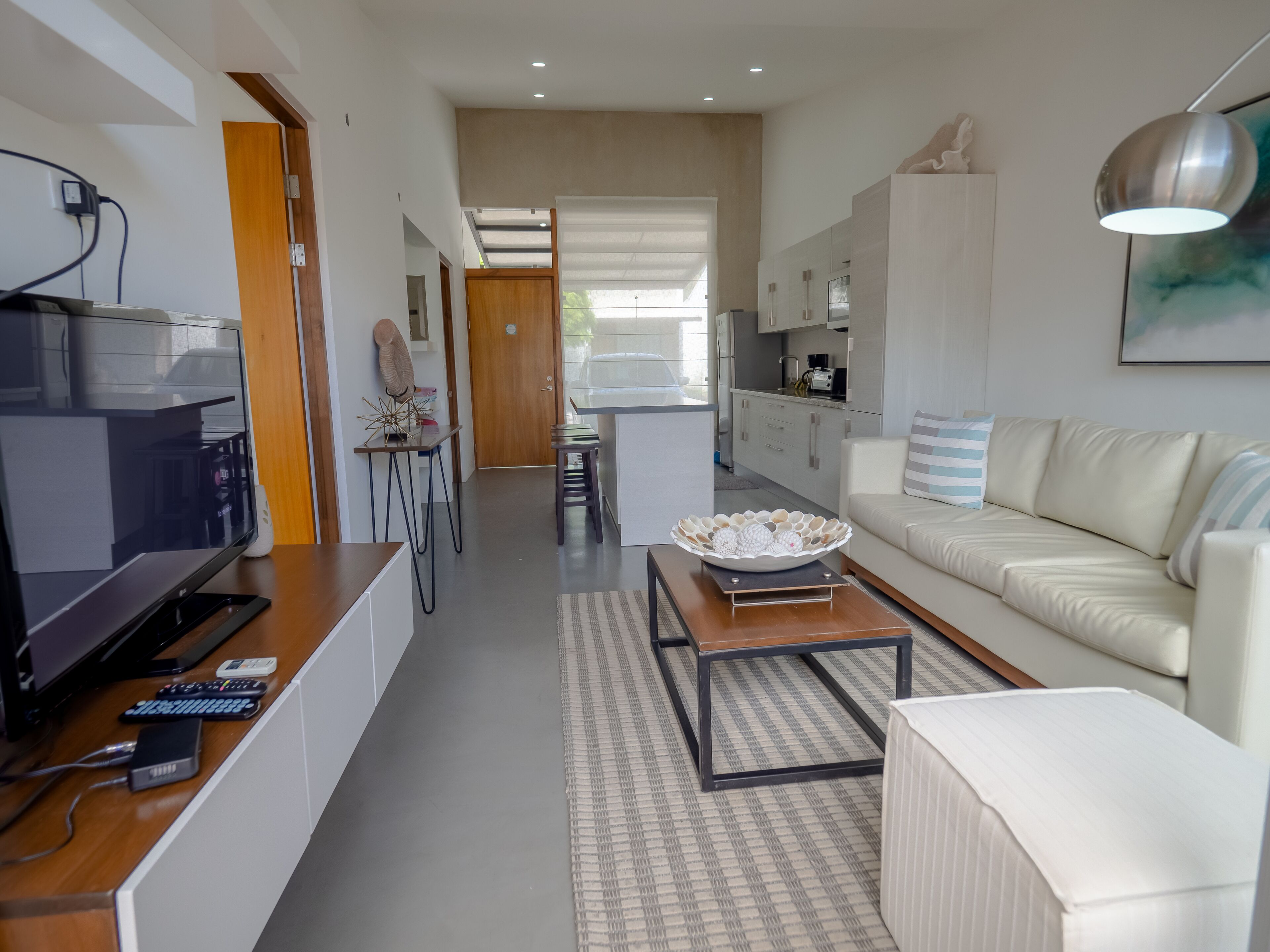 A modern living room with a flat-screen TV, a coffee table, and a sofa. There is a kitchen area with cabinets and a dining table.