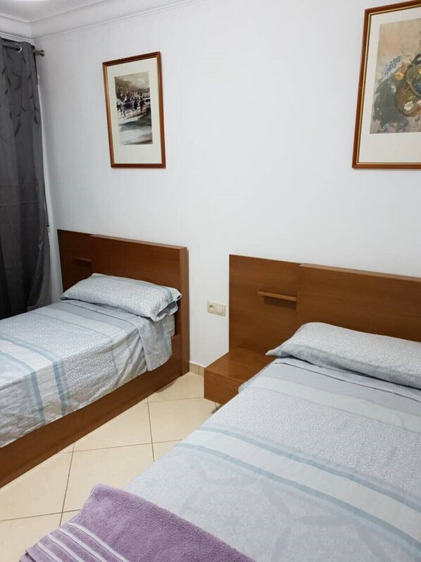 Een kamer met twee eenpersoonsbedden, een klein nachtkastje, een lijst met een foto aan de muur en een gordijn.