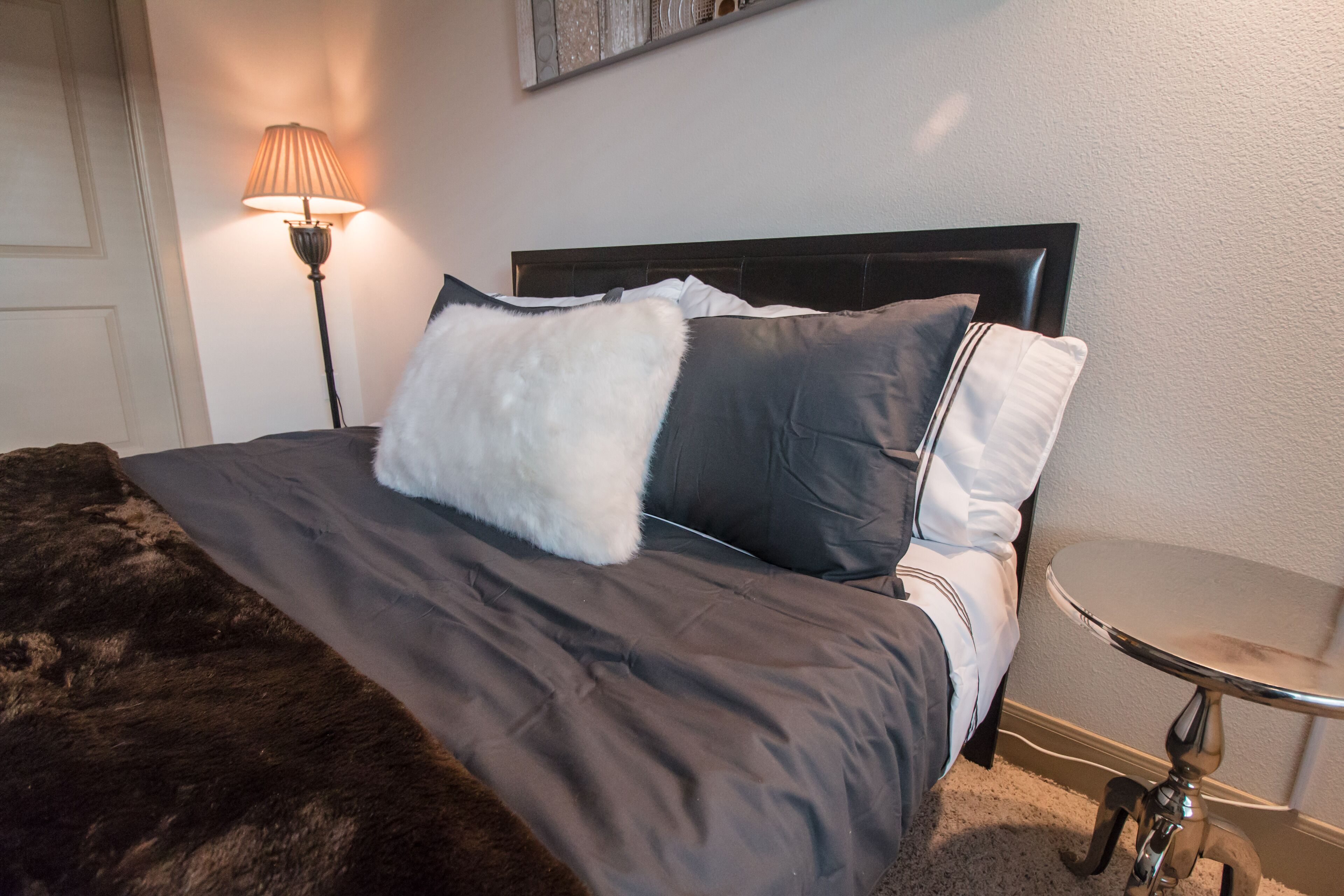 A neatly made bed with a dark brown blanket and two pillows, a small round table beside it, and a floor lamp in the corner.