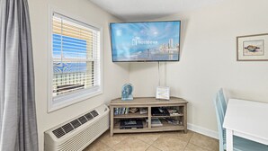 A hotel room with a television mounted on the wall, a window with blinds, a small wooden table, and a chair. - Emerald Skye 54 (Orange Beach)