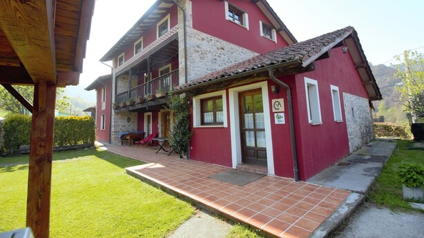 A two-story building with a red facade and a stone base, a balcony, and a small garden area. - TREASURE HOUSE IN RESERVE OF THE BIOSPHERE PICOS DE EUROPA (Cangas de Onís)