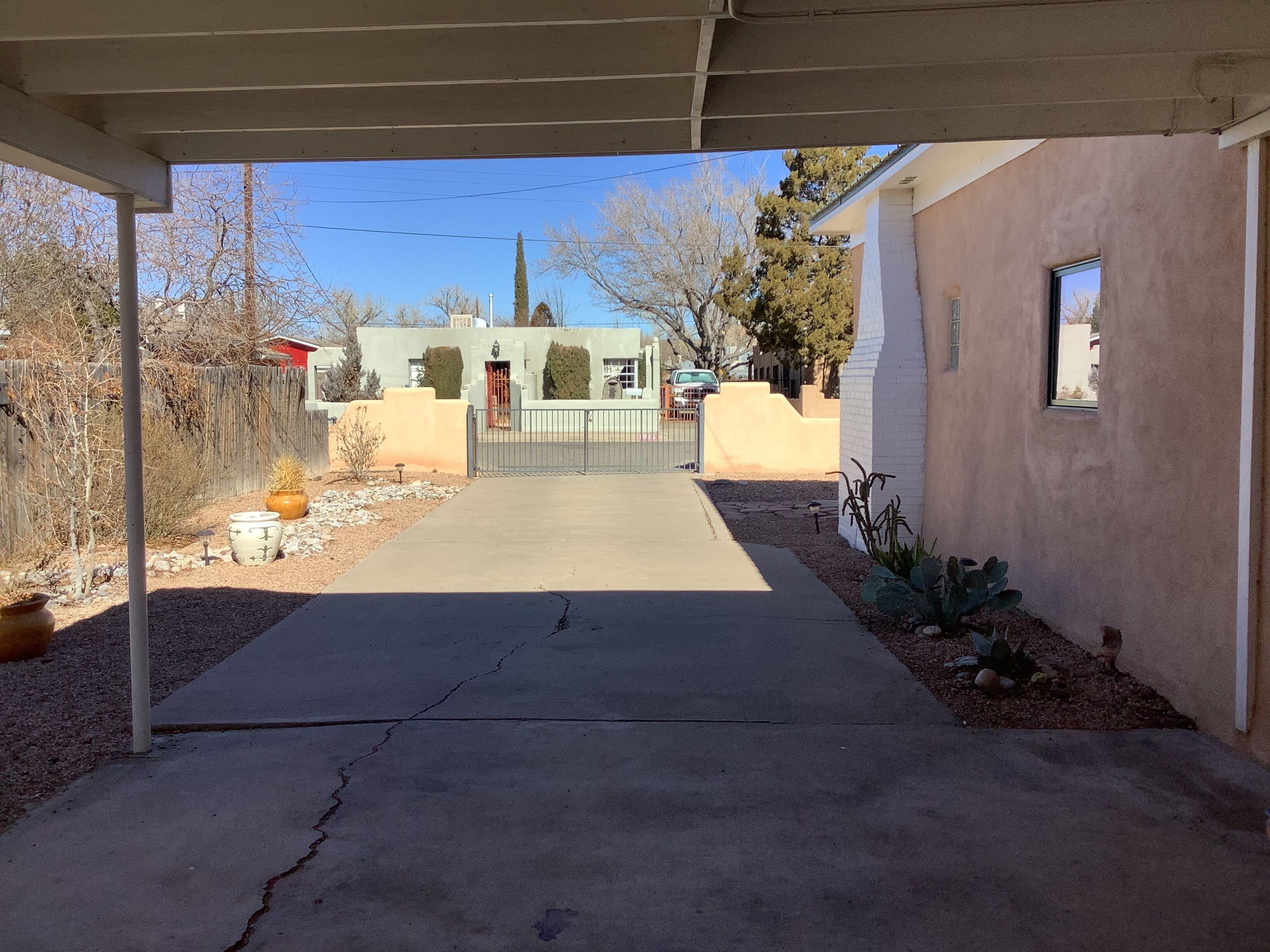 A covered driveway leading to a house with a gated entrance, surrounded by a fence and various plants.
