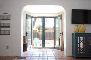 An open doorway leading to a patio area with a blue chair, a TV mounted on the wall, and a cabinet with yellow vases. - Charming Gated Adobe in Historic Old Town  (Albuquerque)