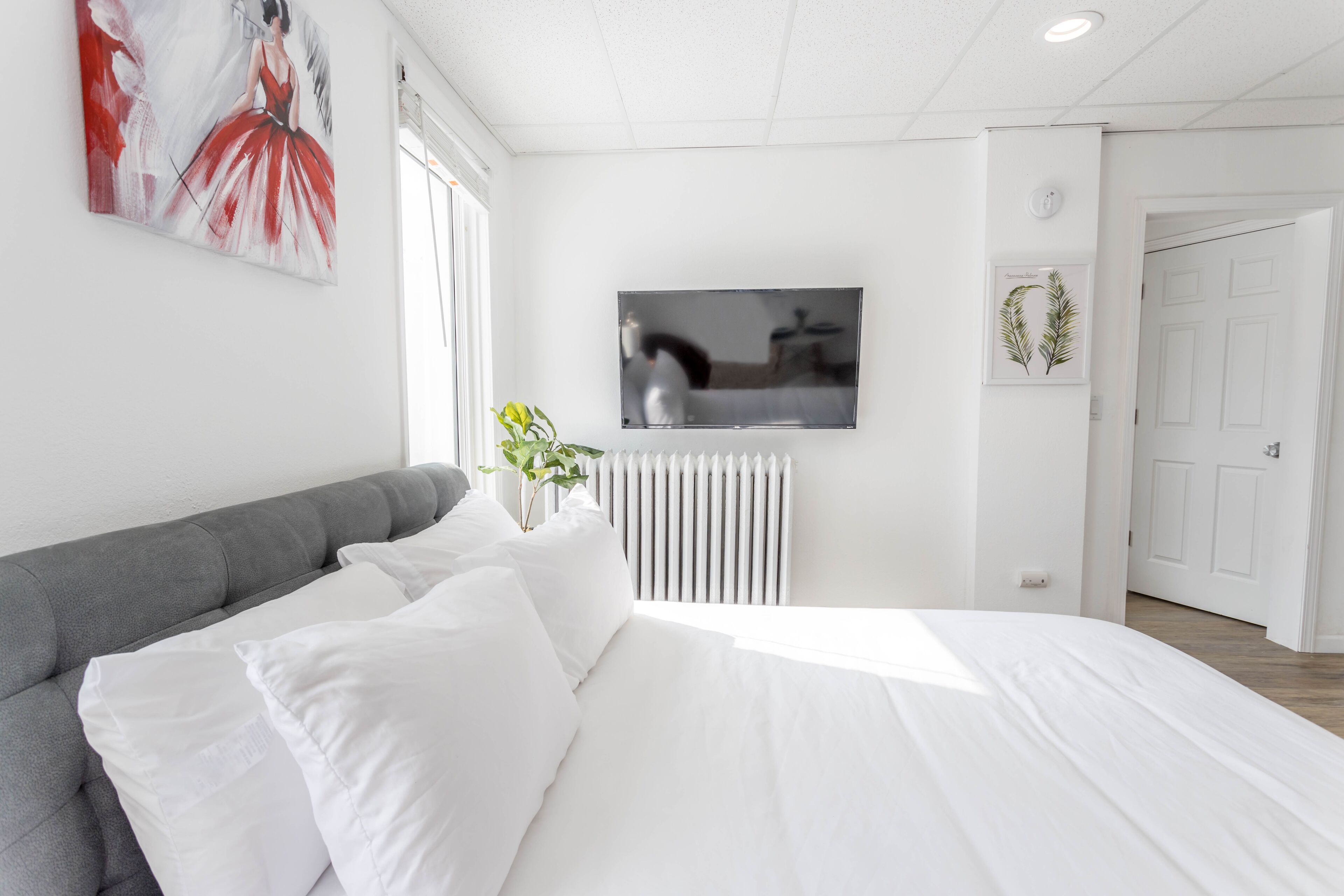 Une chambre moderne avec une tête de lit rembourrée de couleur grise, un grand lit avec du linge de lit blanc, un téléviseur à écran plat fixé au mur et un radiateur.