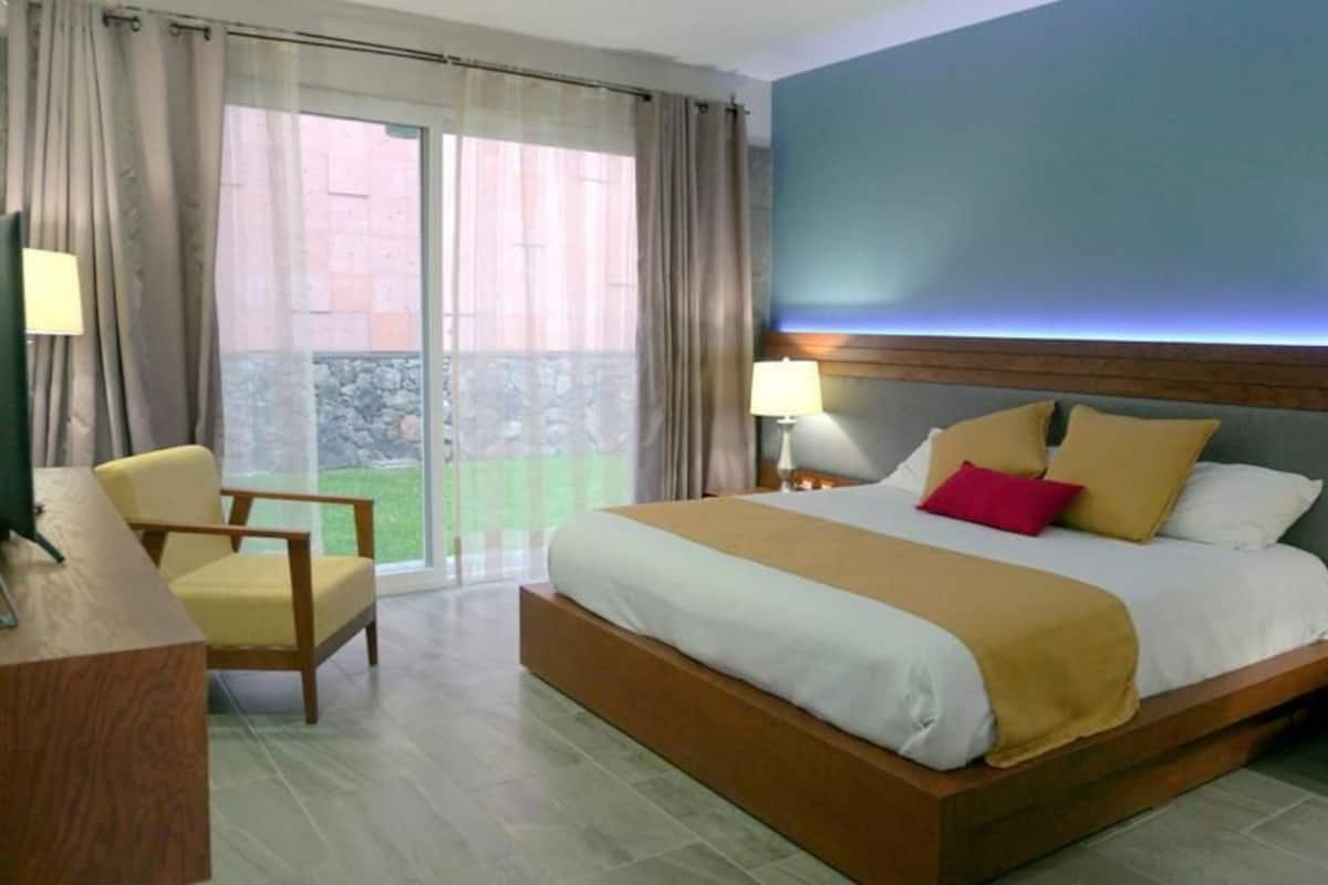 Ein Hotelzimmer mit einem großen Bett, einem Schreibtisch, einem Sessel und Blick auf einen Innenhof durch eine Glastür.