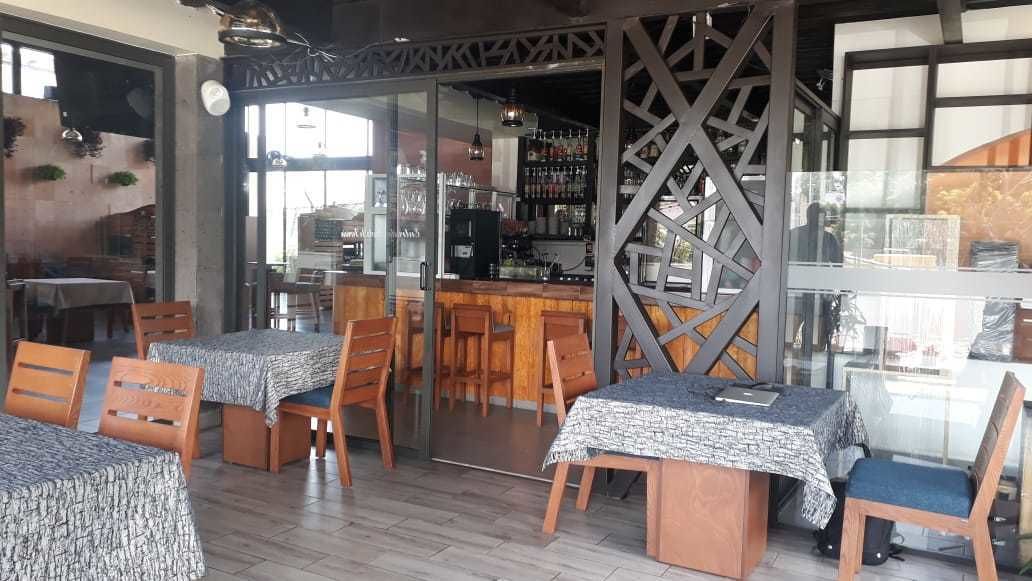 A restaurant with wooden tables and chairs, a bar area, and a kitchen visible through a glass partition.