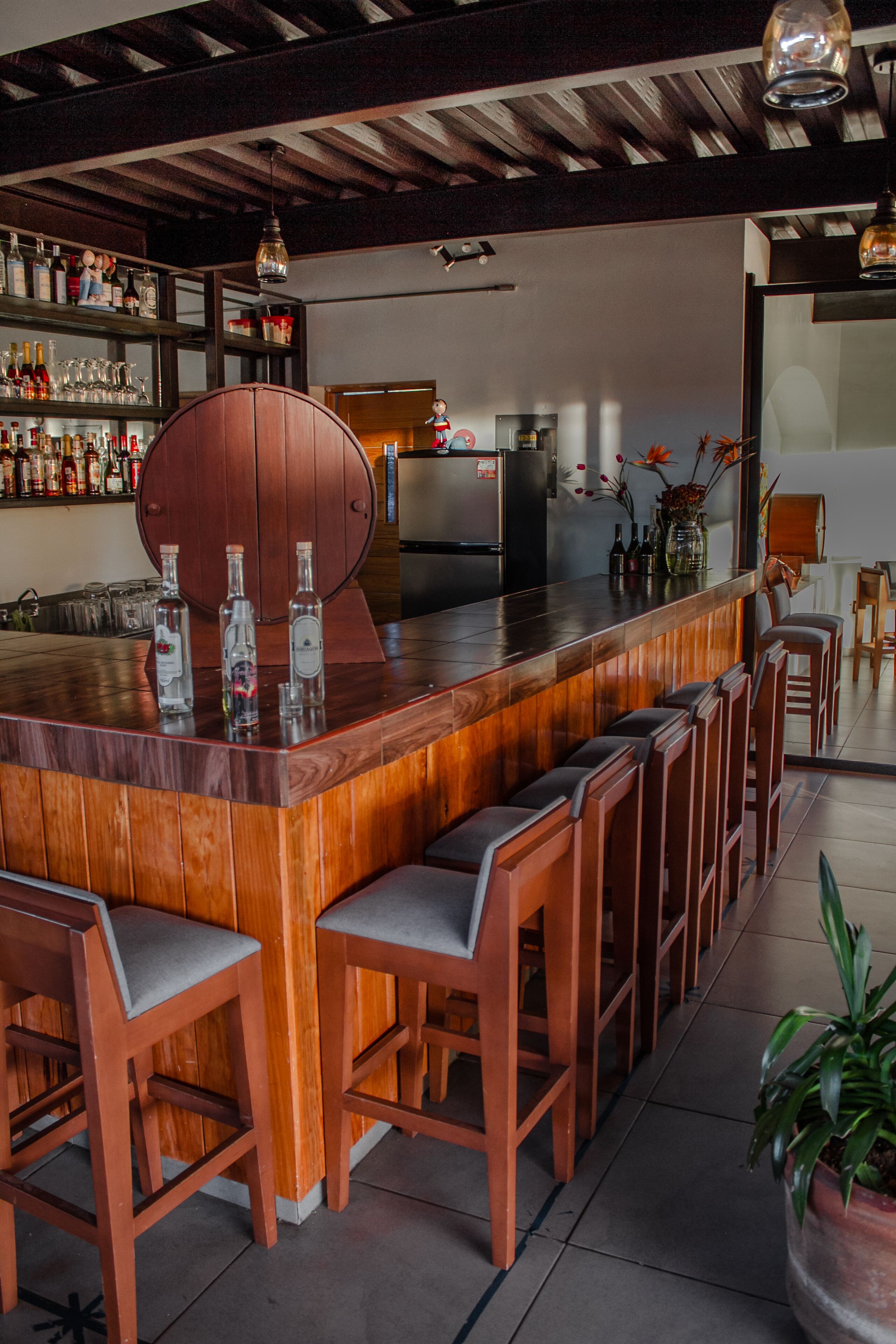Una zona de bar con taburetes de madera, una amplia barra de madera y una variedad de botellas en estanterías.