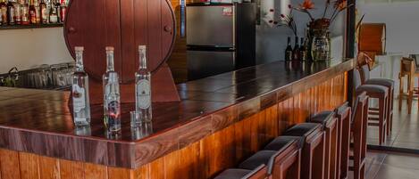 Una zona de bar con taburetes de madera, una amplia barra de madera y una variedad de botellas en estanterías.