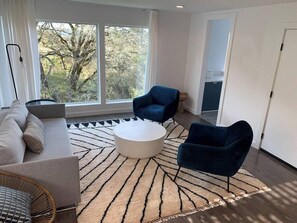 Un salon moderne avec un canapé, deux fauteuils, une table basse et une grande fenêtre donnant sur la verdure.