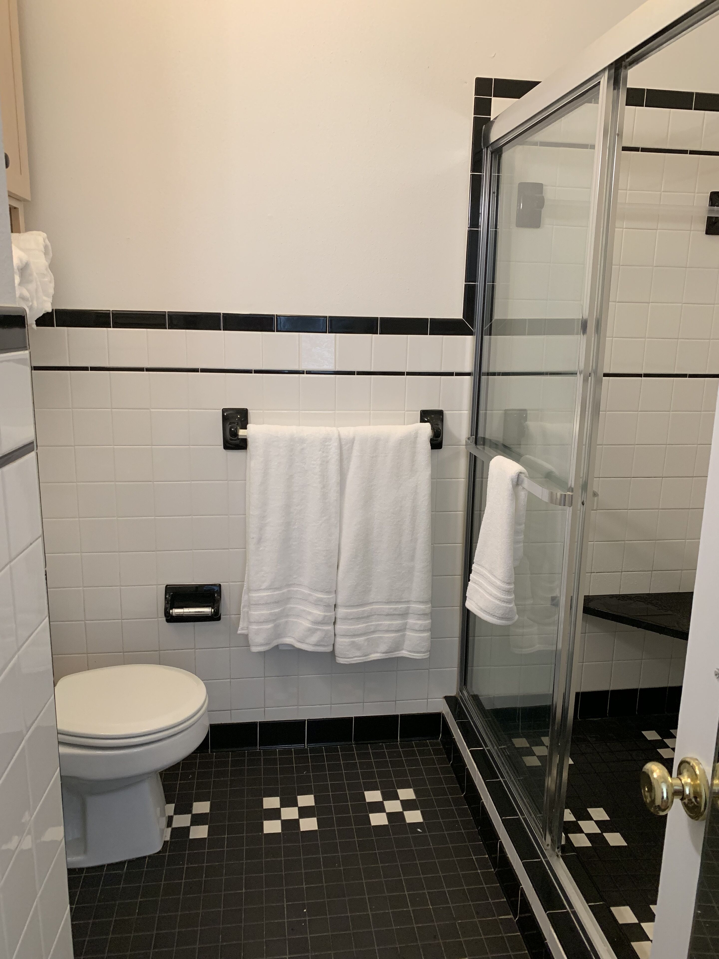 A bathroom with a toilet, two white towels hanging on a rack, a glass shower enclosure, and a black and white checkered floor pattern.