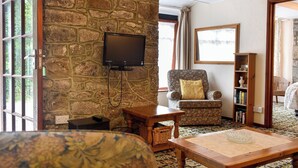 A cozy living room with a stone wall, a television, a floral armchair, a wooden coffee table, and a bookshelf.