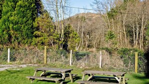 Two picnic tables in a grassy area with a wire fence and trees in the background.