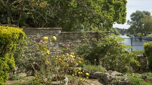 Eine steine Mauer mit einem Garten davor, ein Fluss und eine Brücke im Hintergrund.