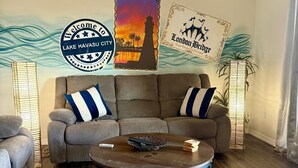 Ein Wohnzimmer mit einem braunen Sofa, zwei blau-weißen Kissen mit Streifen, einem runden Couchtisch mit Zeitschriften und einem Korb, sowie einer Wand mit zwei gerahmten Postern von Lake Havasu City und London Bridge.