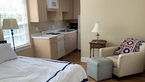 Een slaapkamer met een groot bed, een stoel, een klein tafeltje en een kitchenette op de achtergrond.