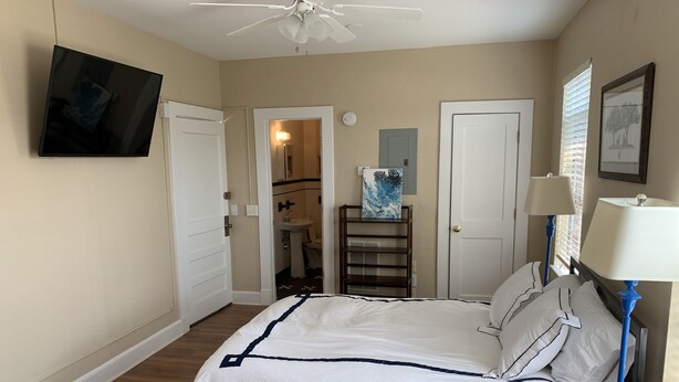 Een slaapkamer met een bed, een aan de muur bevestigde televisie, een plafondventilator en een badkamer door een geopende deur zichtbaar.