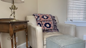 Een kamer met een houten tafel, een witte lamp, een beige bank met een gestreept plaids en een lichtgrijze poef.
