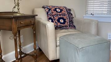 A room with a wooden table, a white lamp, a beige sofa with a patterned throw, and a light grey ottoman.