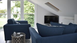A modern living room with a large window, a flat-screen TV, and blue sofas. - 2 bedroom accommodation in St. Lawrence, near Burnham-on-Crouch (St. Lawrence, near Burnham-on-Crouch)