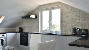 A modern kitchen with a glass dining table, white chairs, a built-in oven, and a window with a view of greenery. - 2 bedroom accommodation in St. Lawrence, near Burnham-on-Crouch (St. Lawrence, near Burnham-on-Crouch)