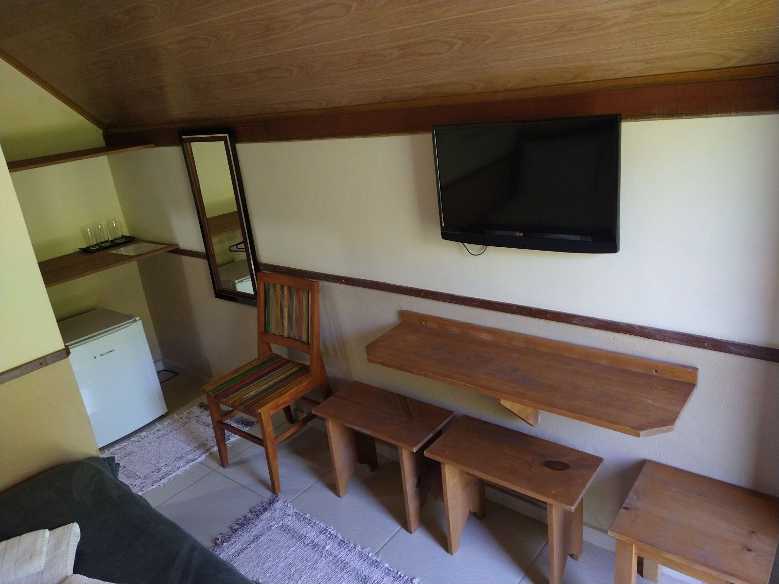 A small, cozy room with a wooden ceiling, a television mounted on the wall, a wooden chair, a small table, and a bench.