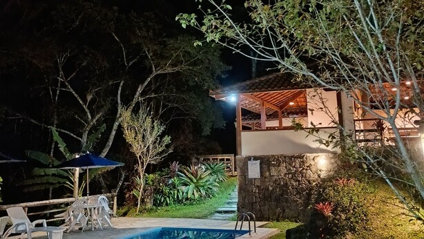 A nighttime view of a house with a swimming pool, surrounded by greenery and a stone wall. - Pousada dos Anjos (Nova Friburgo)