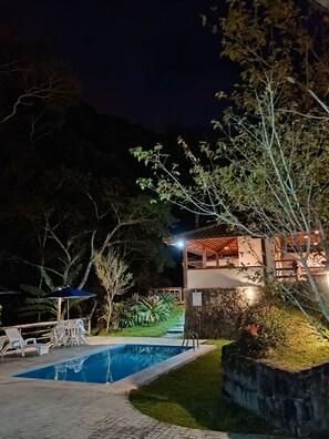 Vista nocturna de una casa con piscina, rodeada de follaje y un muro de piedra.