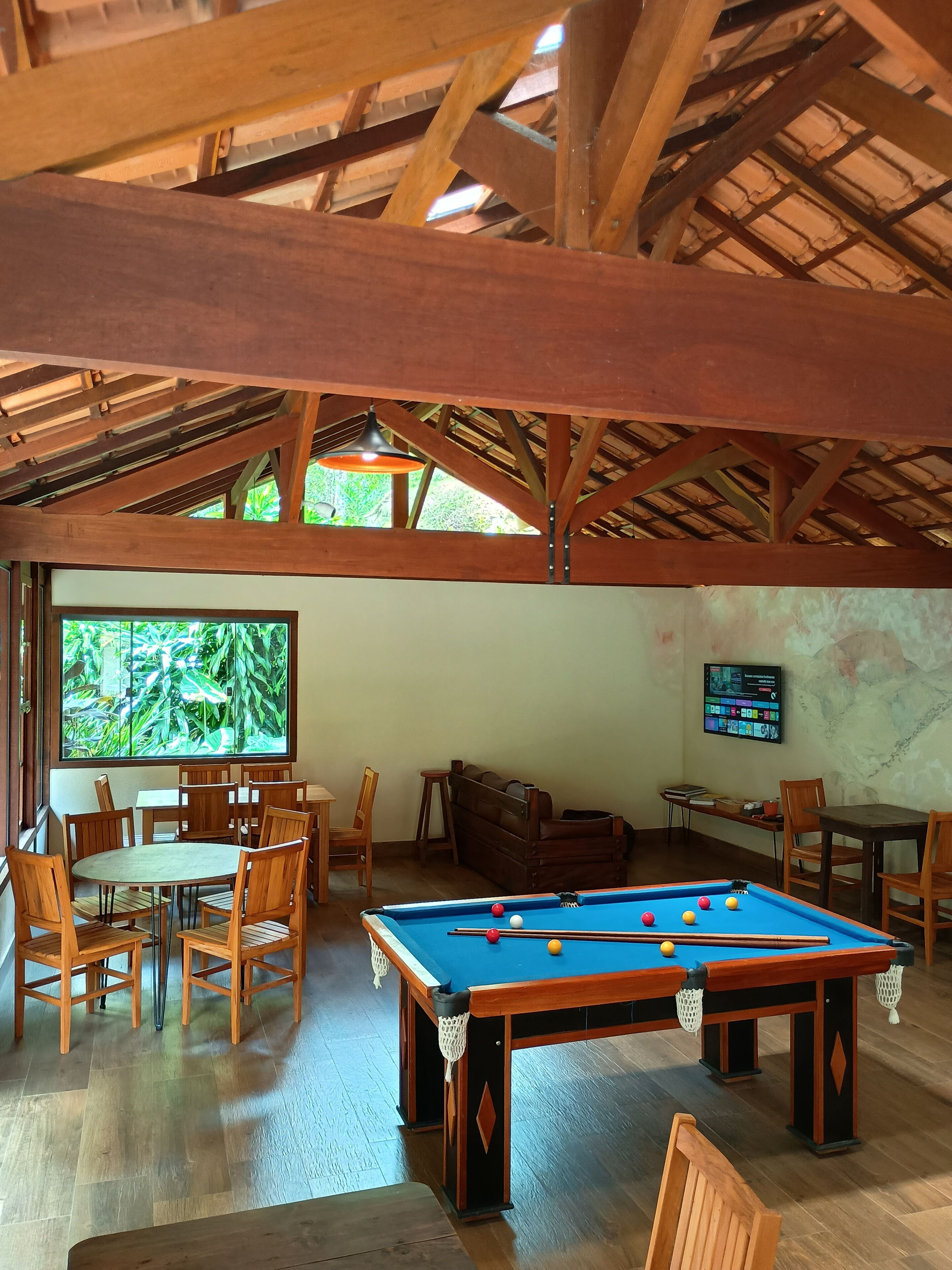 A pool table with chairs, a sofa, and a TV in a room with wooden beams and a large window.