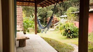 A covered patio with a hammock, a stone bench, and a garden with various plants and a red wall. - Pousada dos Anjos (Nova Friburgo)