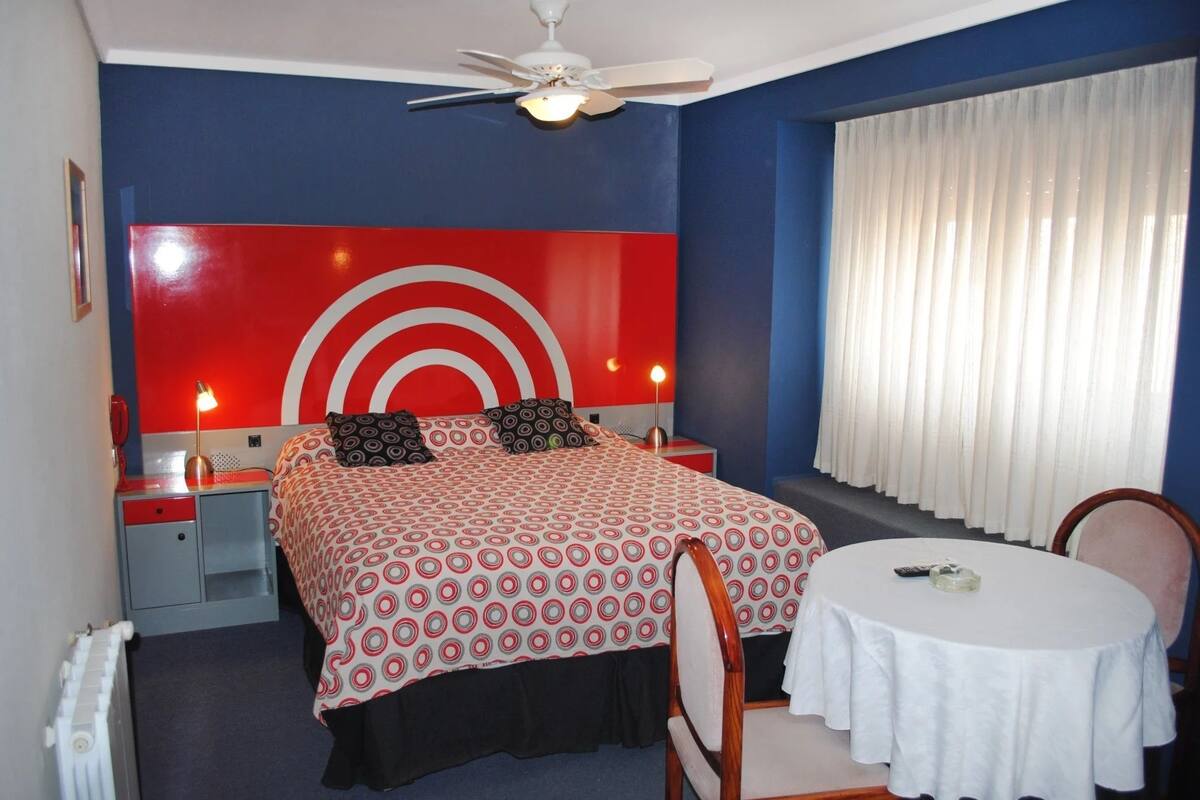 Une chambre d’hôtel comprenant un lit, des tables de chevet, une table ronde, une chaise, un ventilateur de plafond et une fenêtre avec des rideaux.