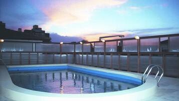 Una piscina en la azotea con barandilla de vidrio y pasamanos de metal, rodeada por un paisaje urbano al atardecer.