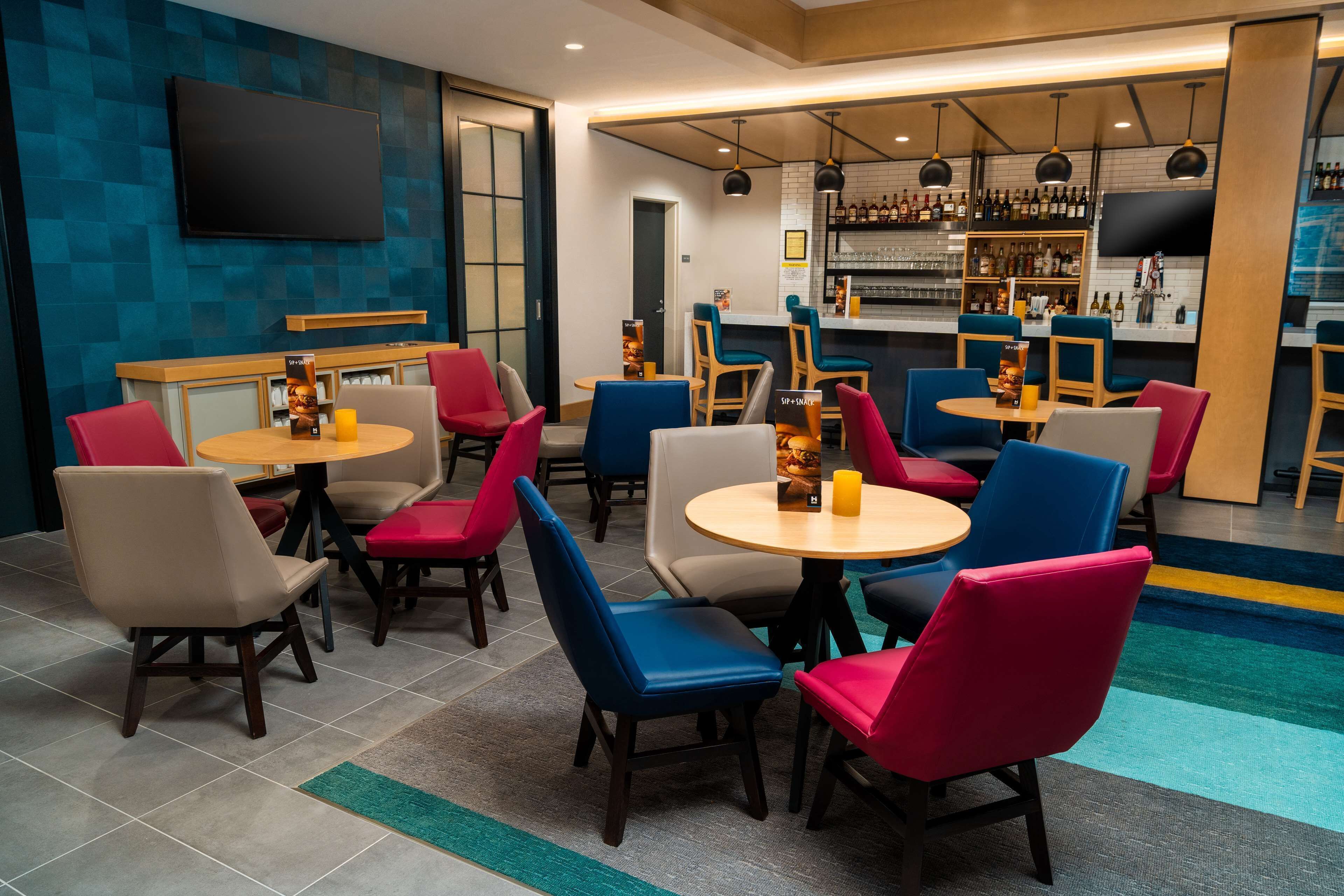 A modern bar area with round tables and colorful chairs, a bar counter with bottles, and a flat-screen TV mounted on the wall.