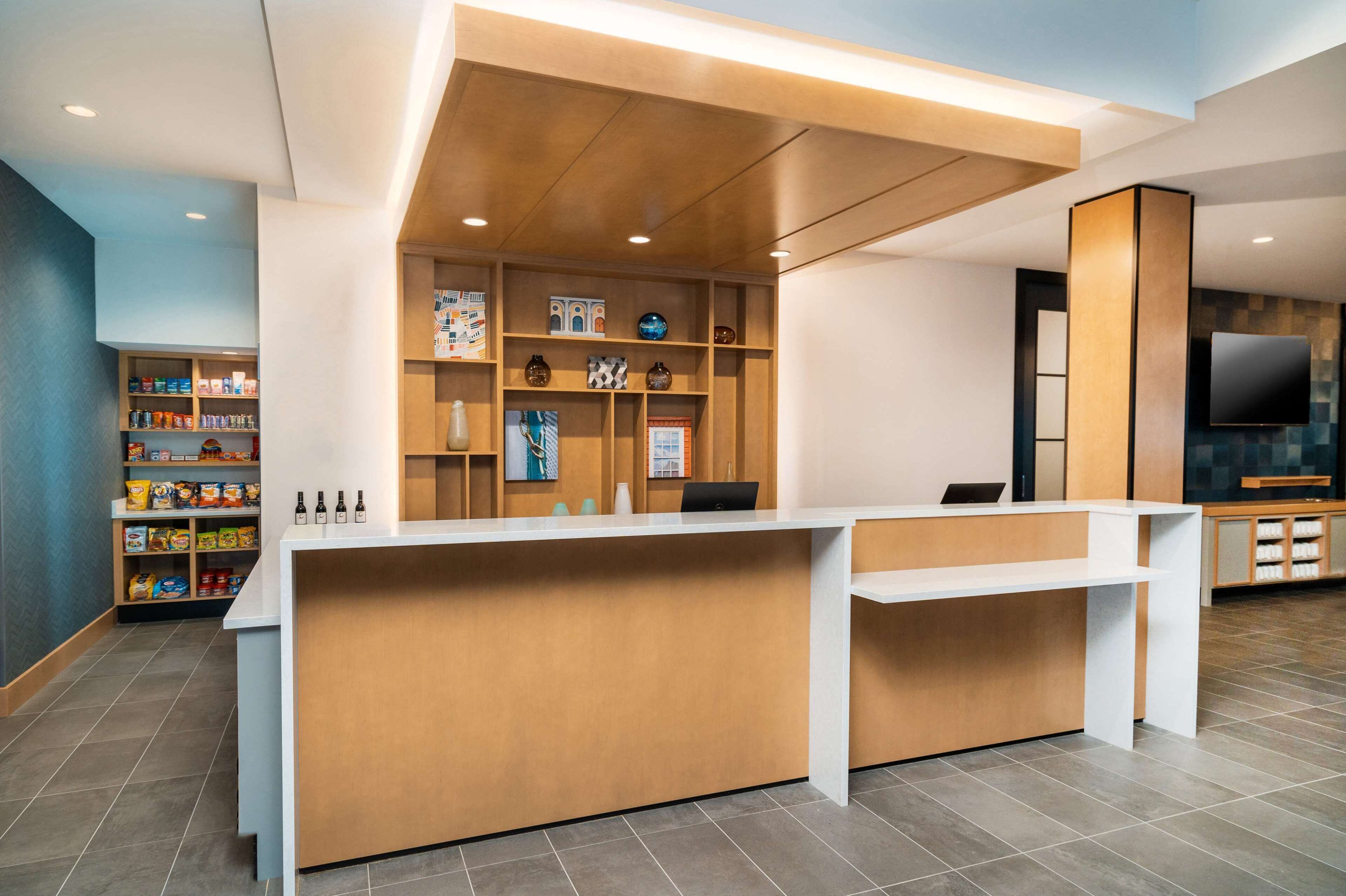 A reception desk with a wooden finish, a white counter, and shelves with various items. There is a television mounted on the wall and a shelf with snacks.