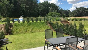 Un espace patio aménagé avec une table et des chaises, un barbecue, et une vue sur un jardin paysager avec des arbres et une clôture en arrière-plan.